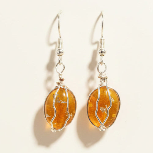 A pair of handmade dangle earrings featuring oval amber-colored glass beads wrapped in silver wire, hanging from silver fishhook ear wires on a brown textured cardboard background.
