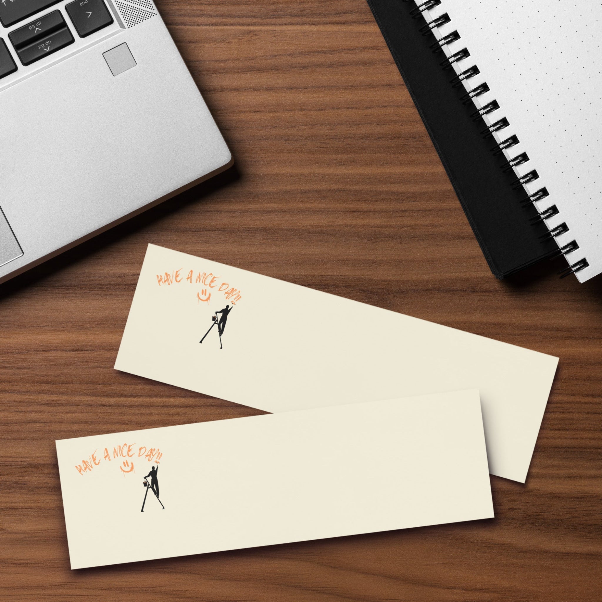 Beige post it sticky note pad with black silhouette and text 'Have a nice day' on a wood desk with a laptop and  spiral notebook