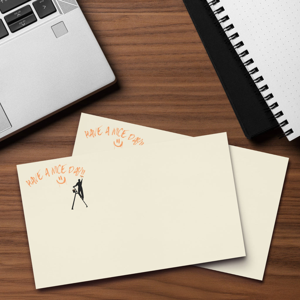 Beige post it sticky note pad with black silhouette and text 'Have a nice day' on a wood desk with a laptop and  spiral notebook