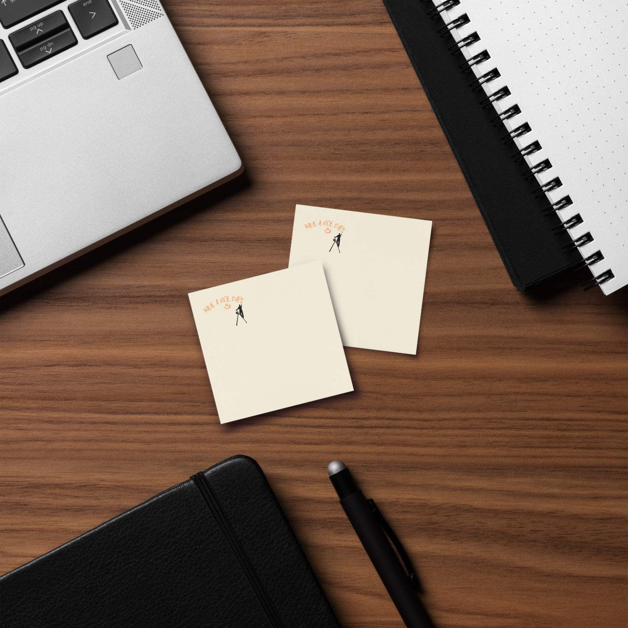 Beige post it sticky note pad with black silhouette and text 'Have a nice day' on a wood desk with a laptop and  spiral notebook
