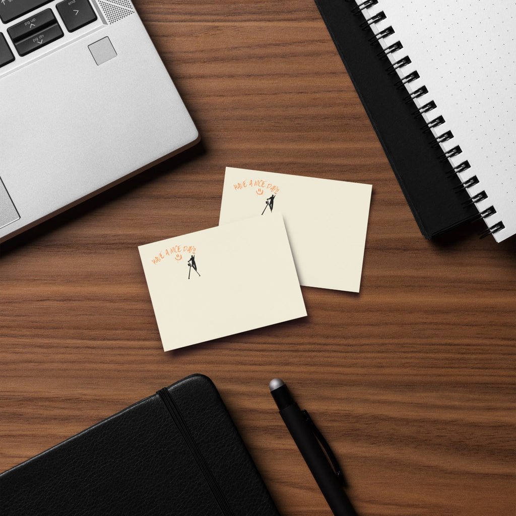 Beige post it sticky note pad with black silhouette and text 'Have a nice day' on a wood desk with a laptop and  spiral notebook
