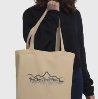 Woman put a tote bag on her shoulder that has a horses and mountains earth tone design on the front of the bag. 