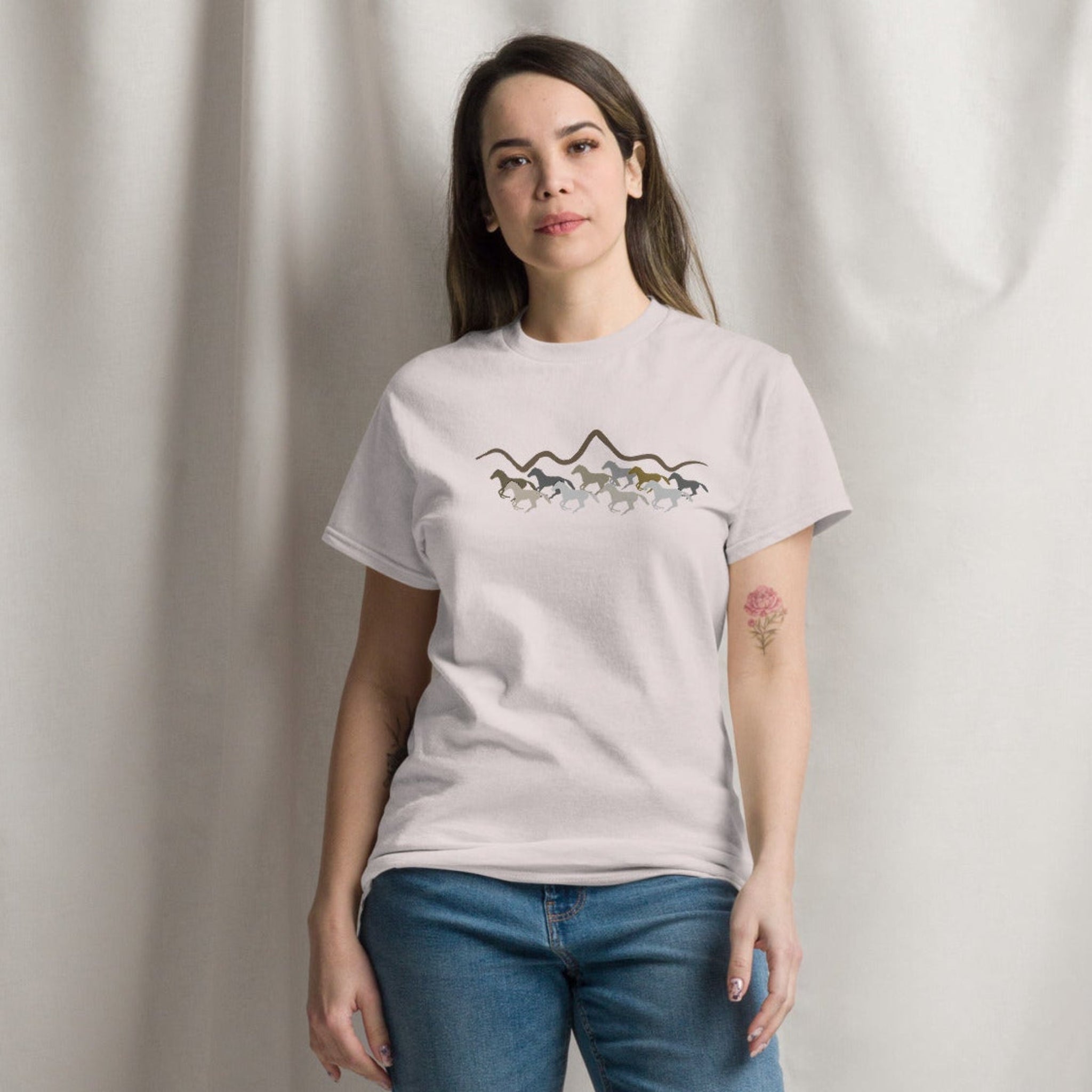Woman wearing a light grey graphic t-shirt featuring a wild mustang stampede and mountain design, styled casually for a western boho look
