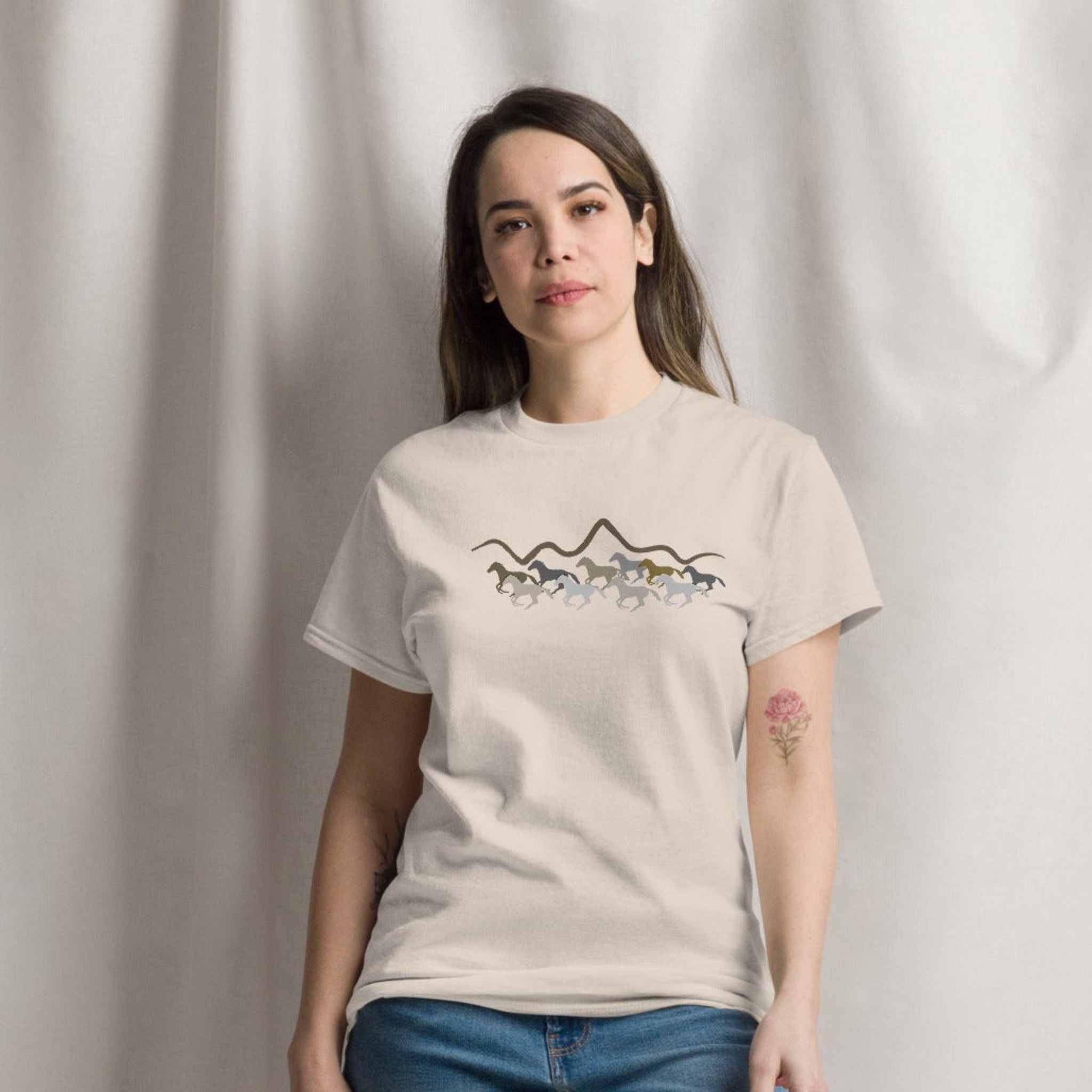 Woman wearing a sand beige graphic t-shirt featuring a wild mustang stampede and mountain design, styled casually for a western boho look