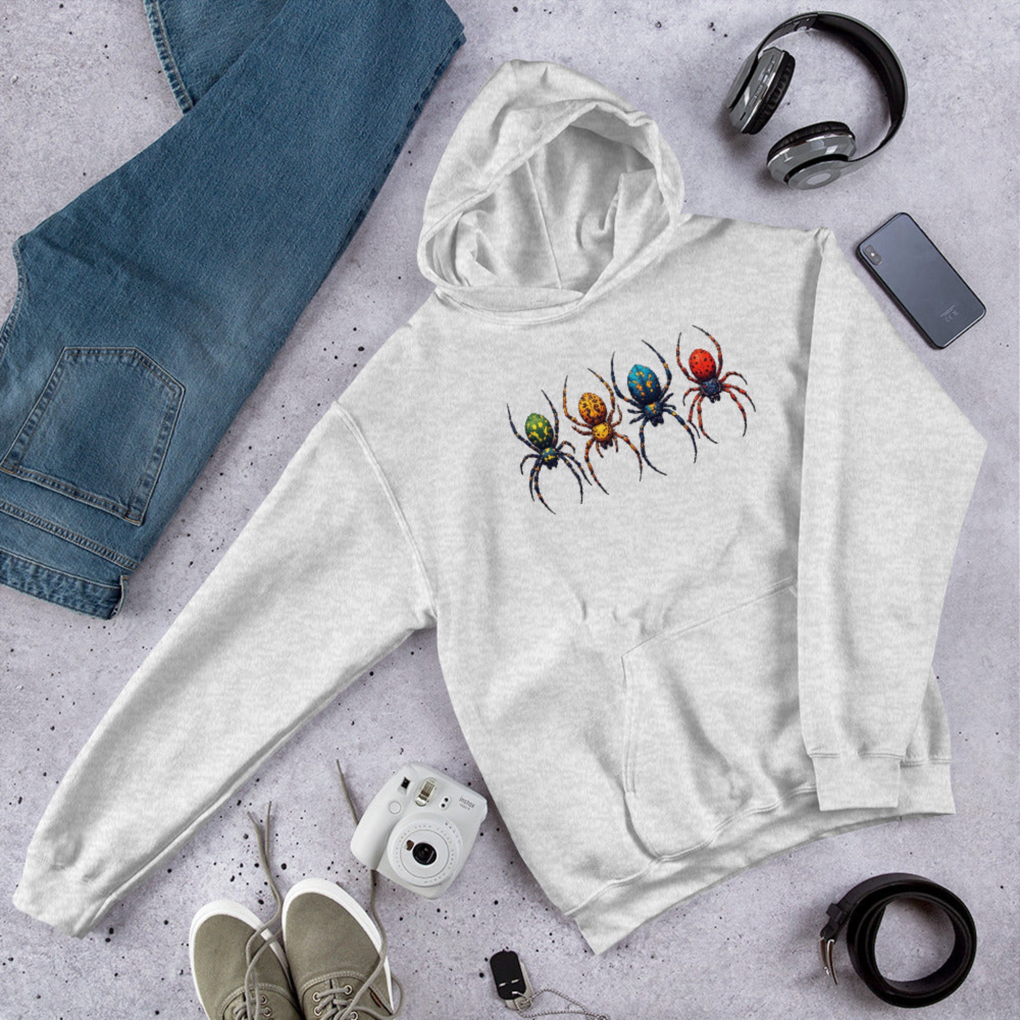 A light heather gray hoodie laid flat on a textured gray surface, featuring a graphic of four colorful spiders on the chest. The hoodie is styled in a flat lay with blue jeans, gray headphones, a smartphone, a white instant camera, beige sneakers, and a black belt.