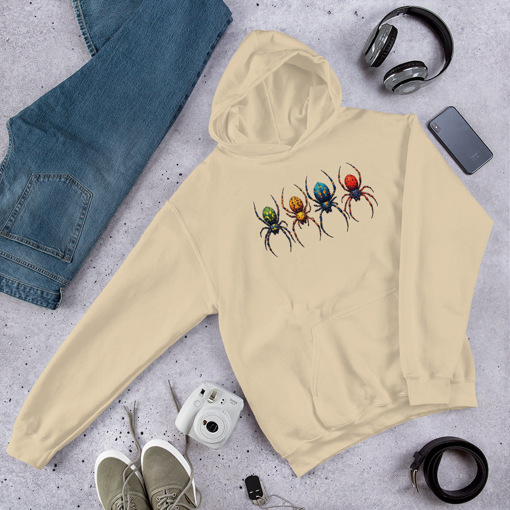 A beige hoodie laid flat on a light gray textured surface, featuring a graphic of four colorful spiders on the chest. The hoodie is styled in a flat lay with blue jeans, black headphones, a smartphone, a white instant camera, gray sneakers, and a black belt.