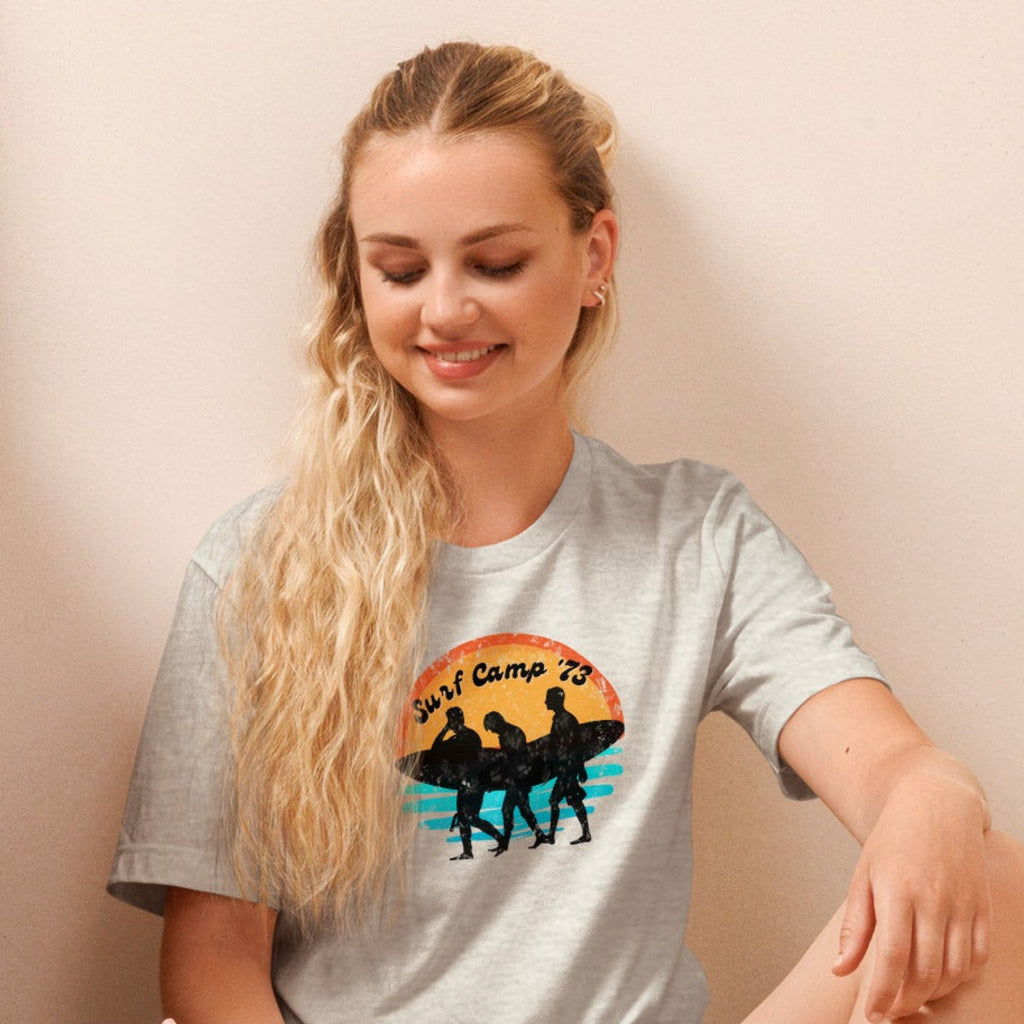 Woman sitting on the floor reading a book wearing a t-shirt with 'Surf Camp 73' distressed vintage design on front of t-shirt.