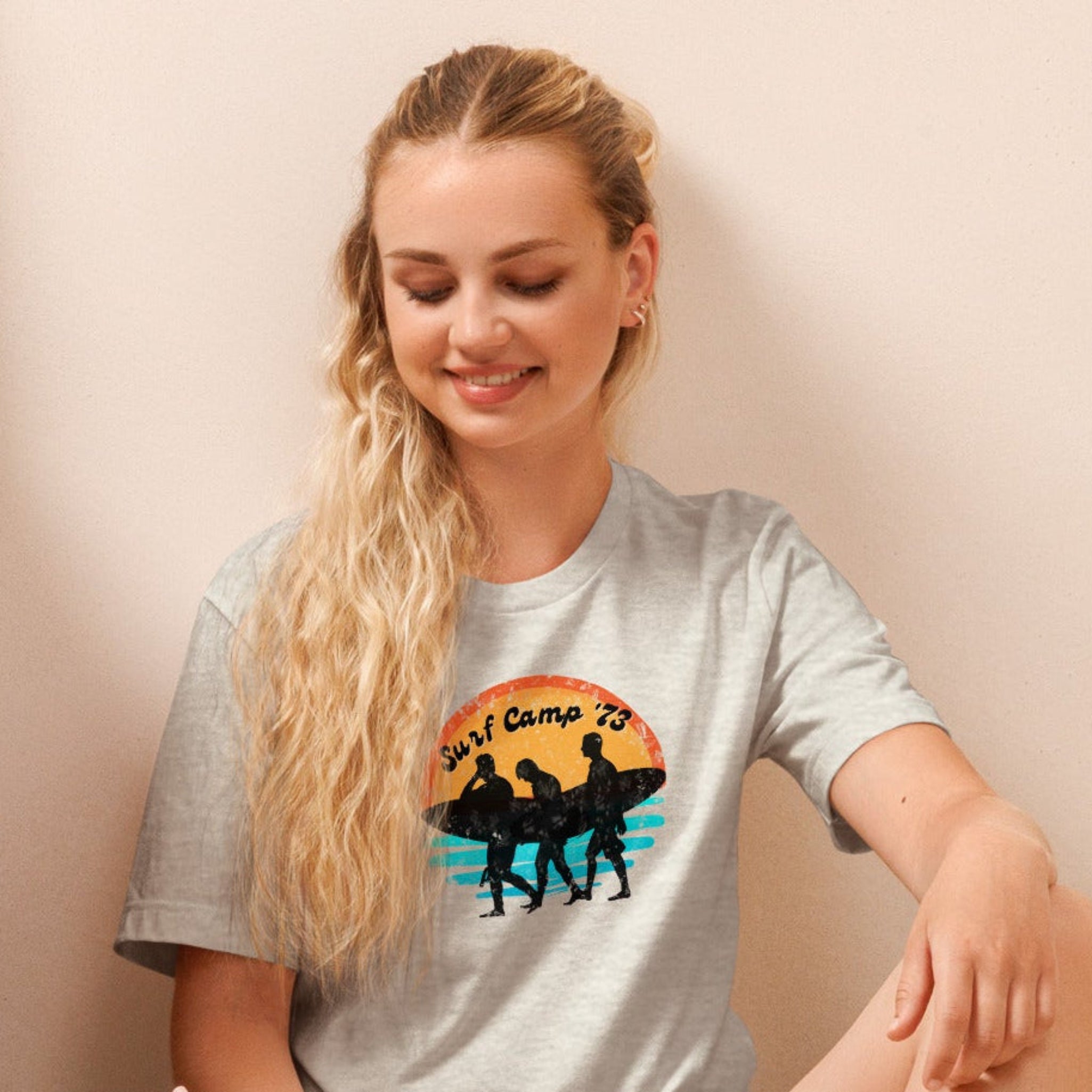 Woman sitting on the floor reading a book wearing a t-shirt with 'Surf Camp 73' distressed vintage design on front of t-shirt.