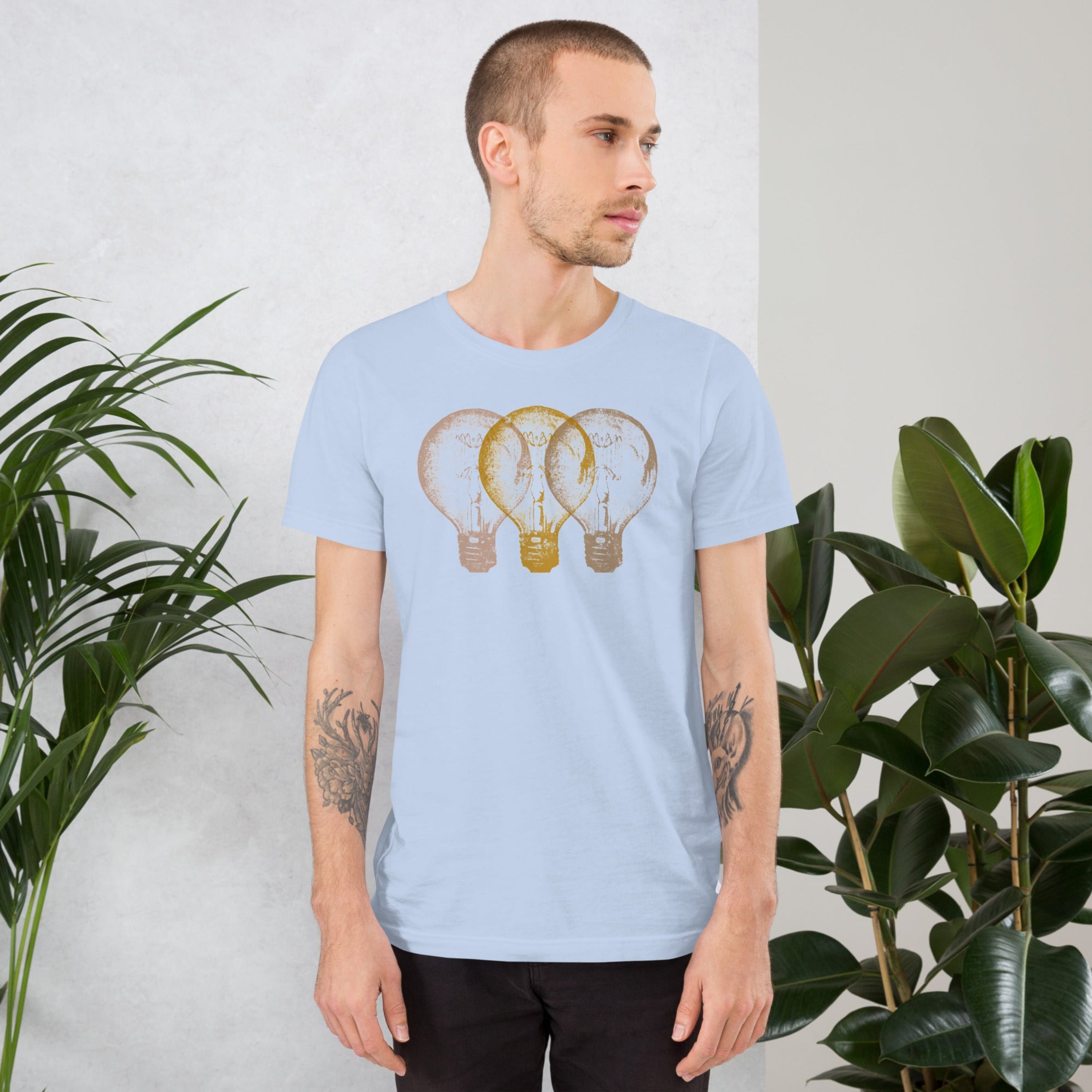 Man wearing a light blue t-shirt with an Edison light bulb graphic design, standing against a white wall with plants.