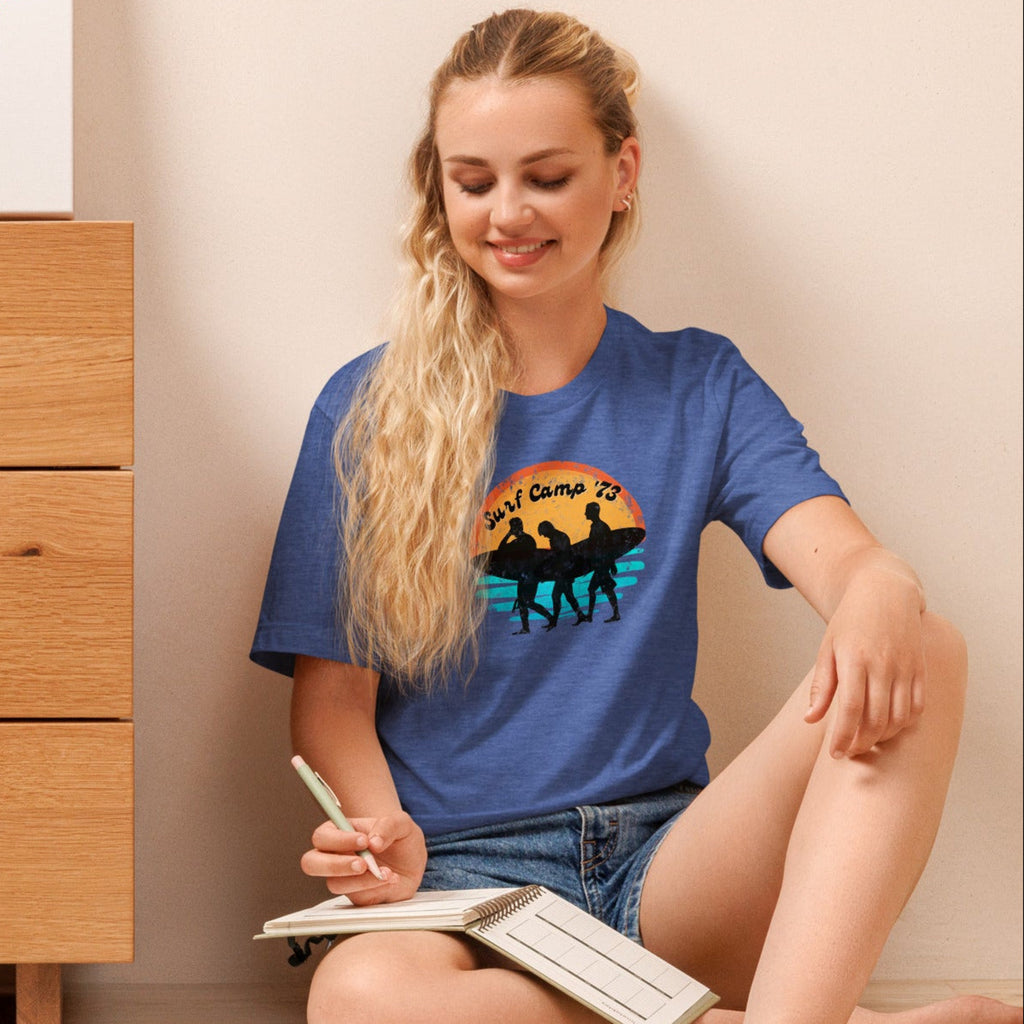 Woman sitting on the floor reading a book wearing a blue t-shirt with 'Surf Camp 73' distressed vintage design on front of t-shirt.