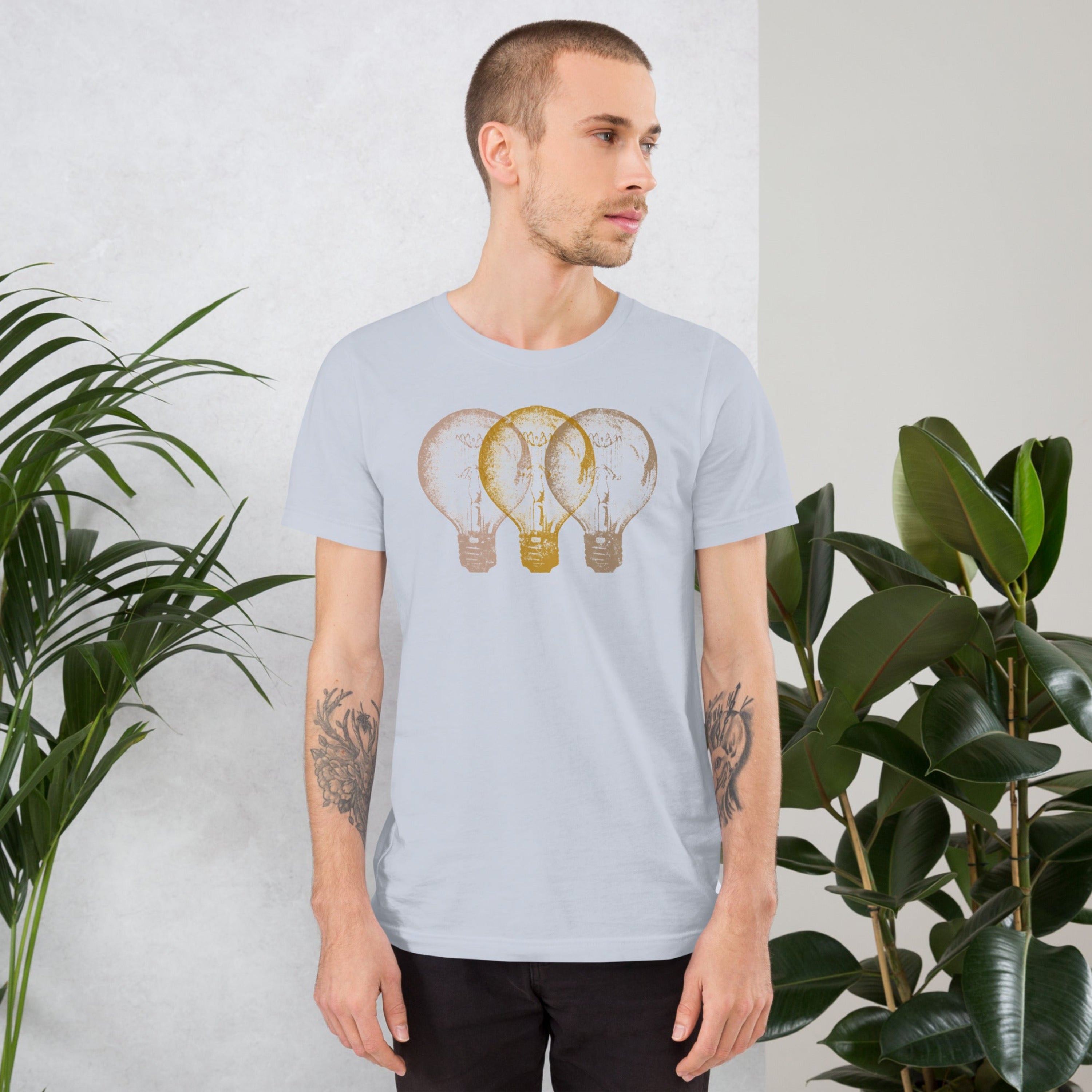 Man wearing a light blue t-shirt with an Edison light bulb graphic design, standing against a white wall with plants.