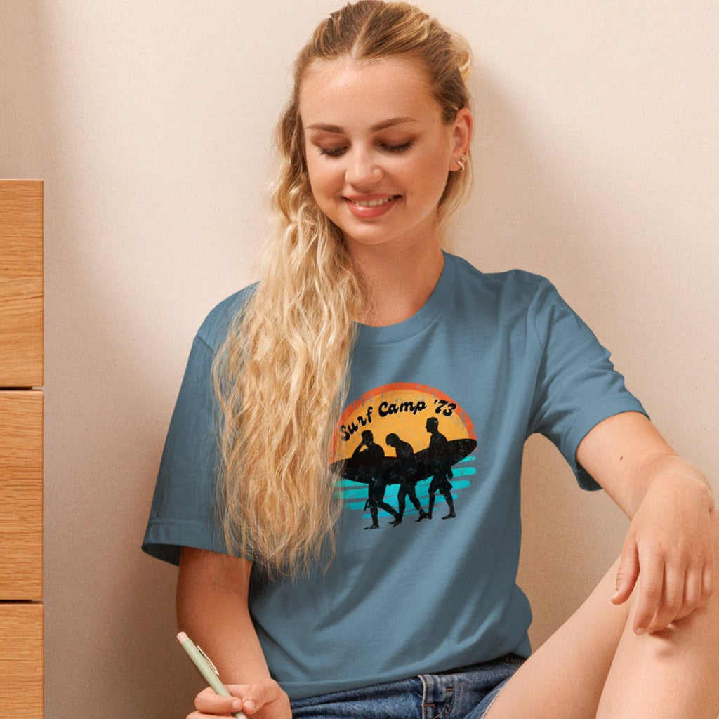 Woman sitting on the floor reading a book wearing a t-shirt with 'Surf Camp 73' distressed vintage design on front of t-shirt.