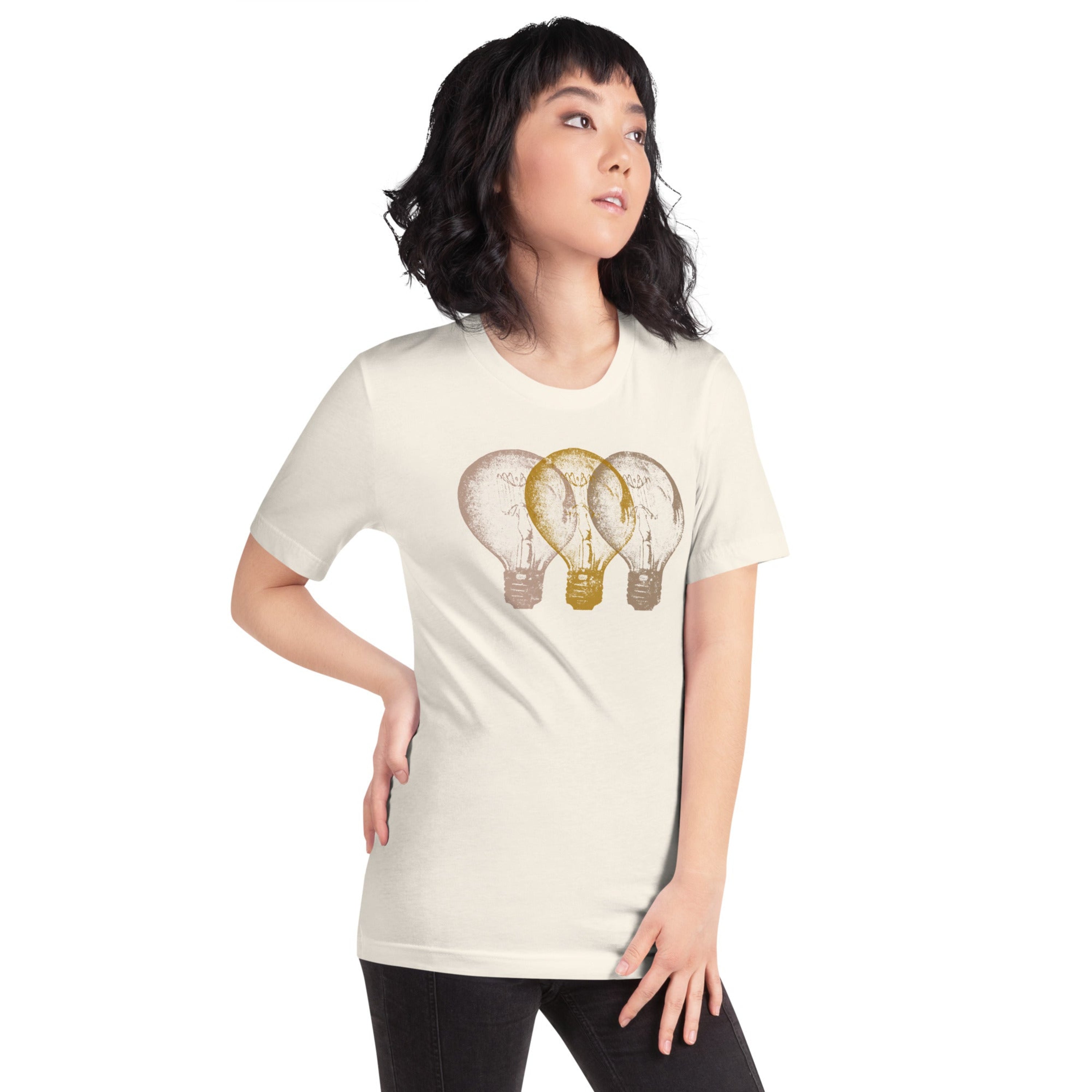 Woman wearing a light sand t-shirt with an Edison light bulb graphic design, standing against a white wall.