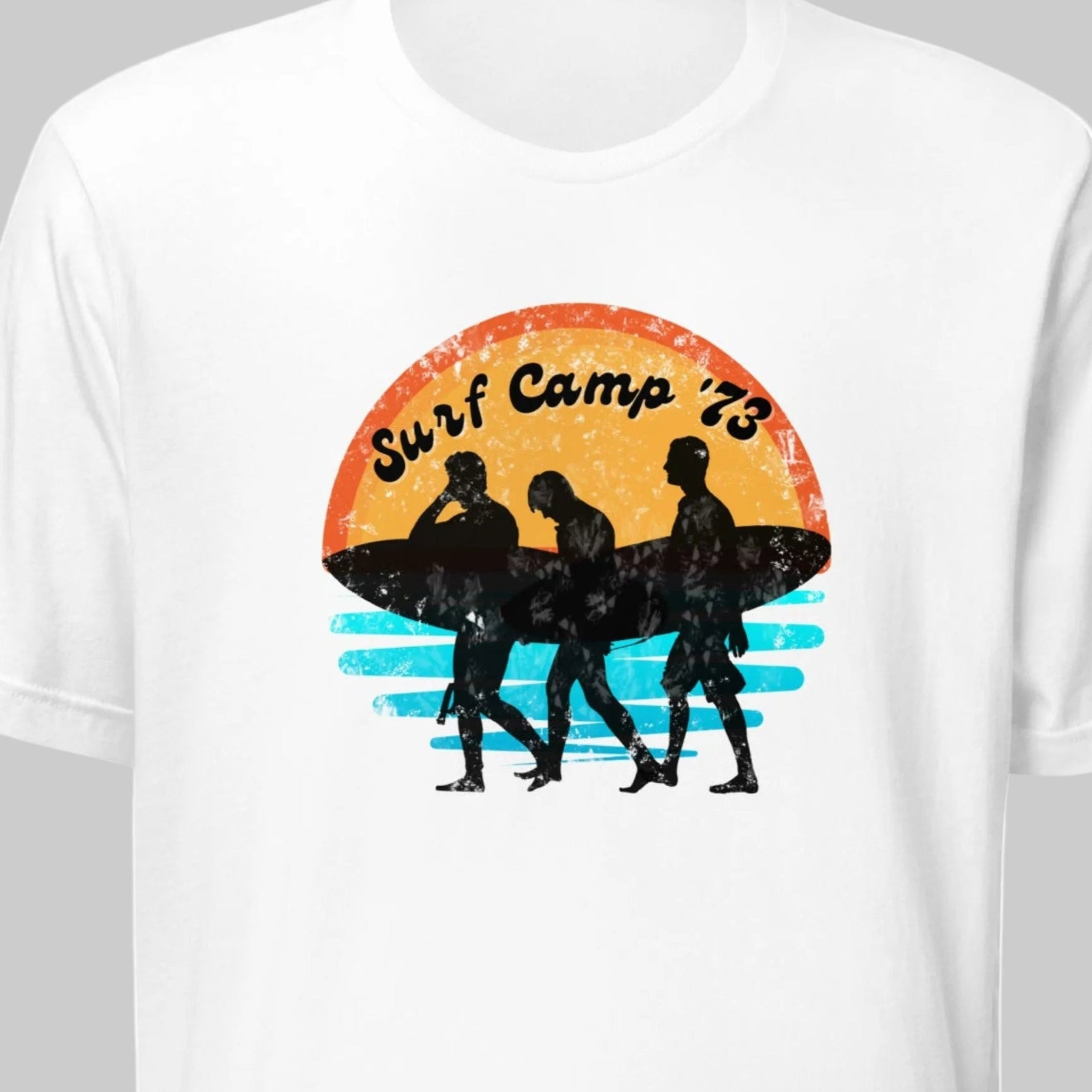 White t-shirt with a graphic design of three surfers and 'Surf Camp 73' text on a grey background.