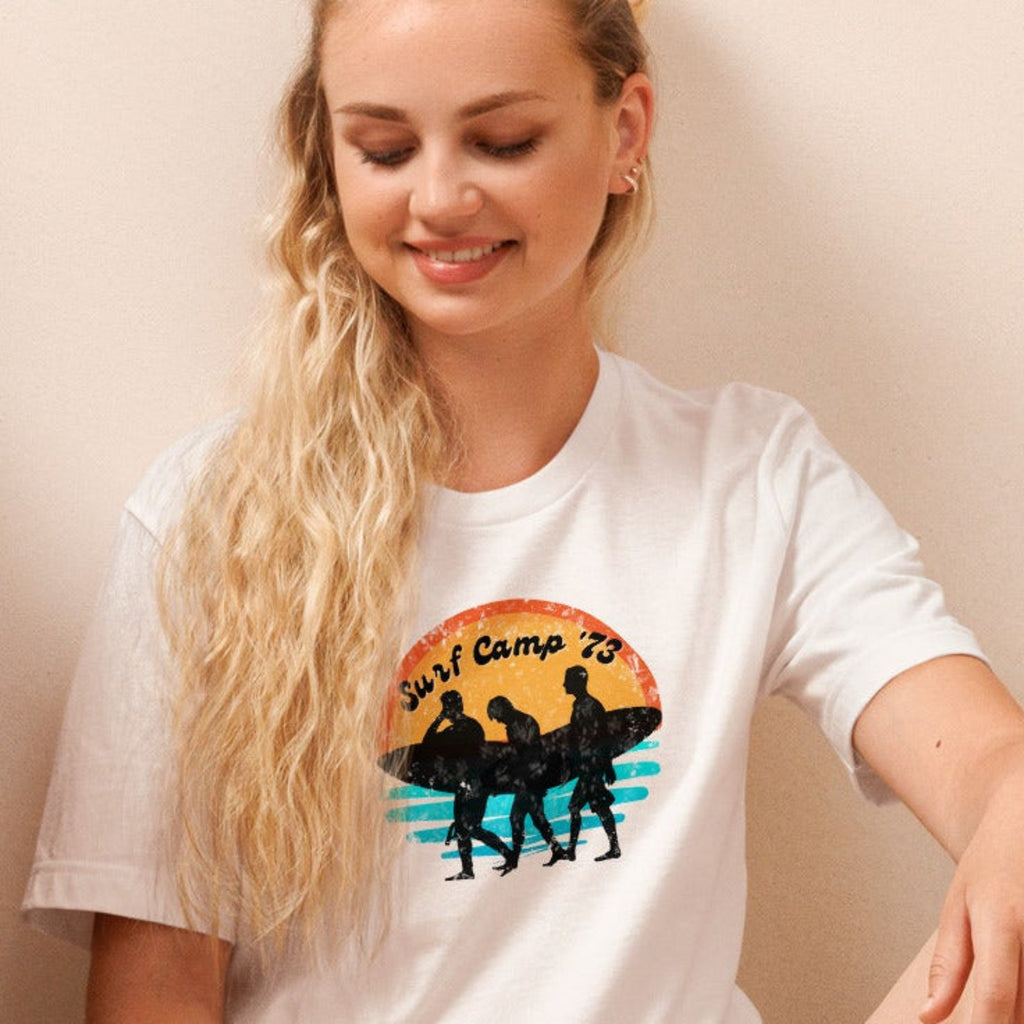 Woman sitting on the floor reading a book wearing a t-shirt with 'Surf Camp 73' distressed vintage design on front of t-shirt.