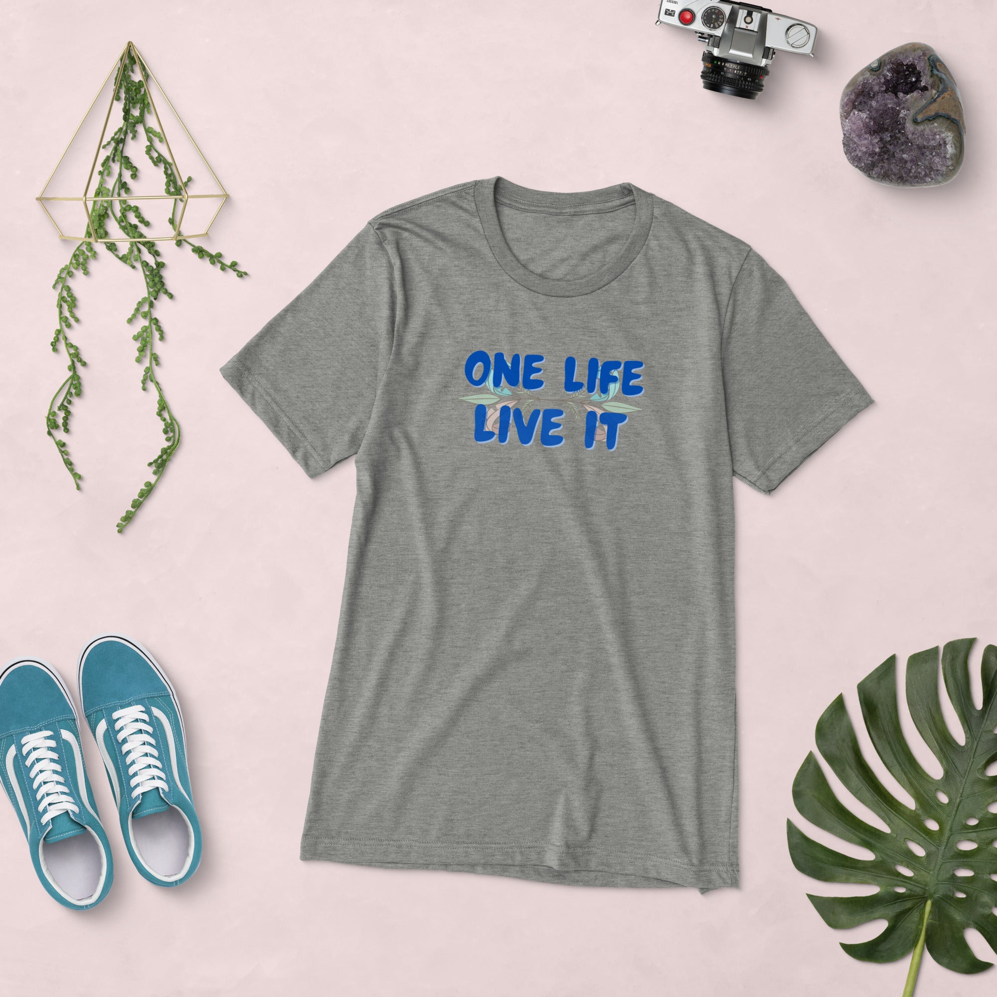 Gray t-shirt with 'ONE LIFE LIVE IT' text, blue sneakers, camera, and plant on a light pink background.