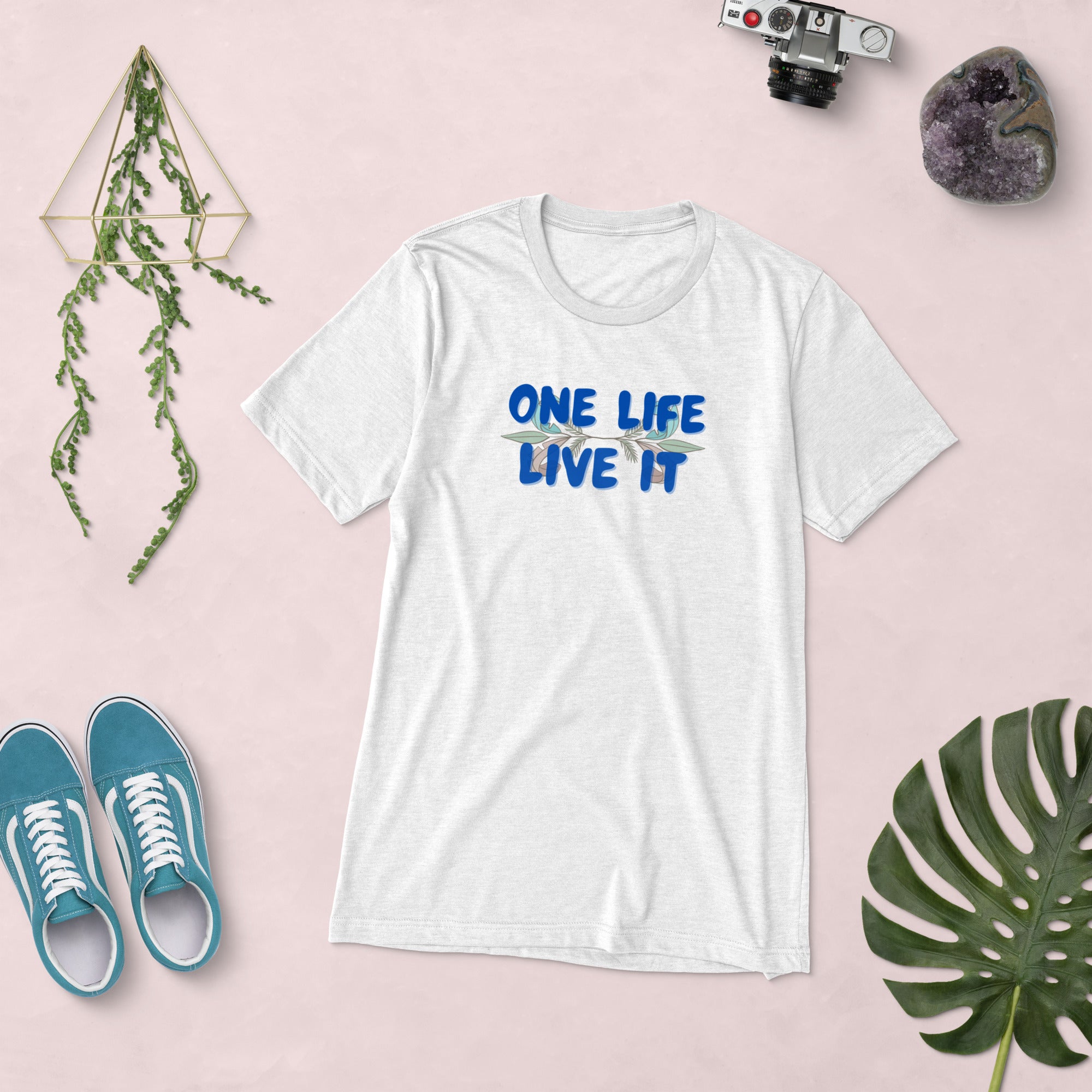 White t-shirt with 'ONE LIFE LIVE IT' text on a pink background with shoes and decor items.
